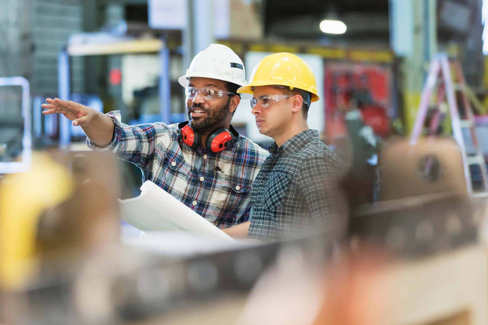 manufacturing engineers wearing safety helmets and glasses reviewing facility plans and discussing production layout on a factory floor