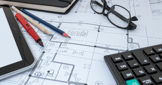 architectural blueprint of a facility layout spread across a desk with a tablet, calculator, eyeglasses, and red and blue pencils placed on top